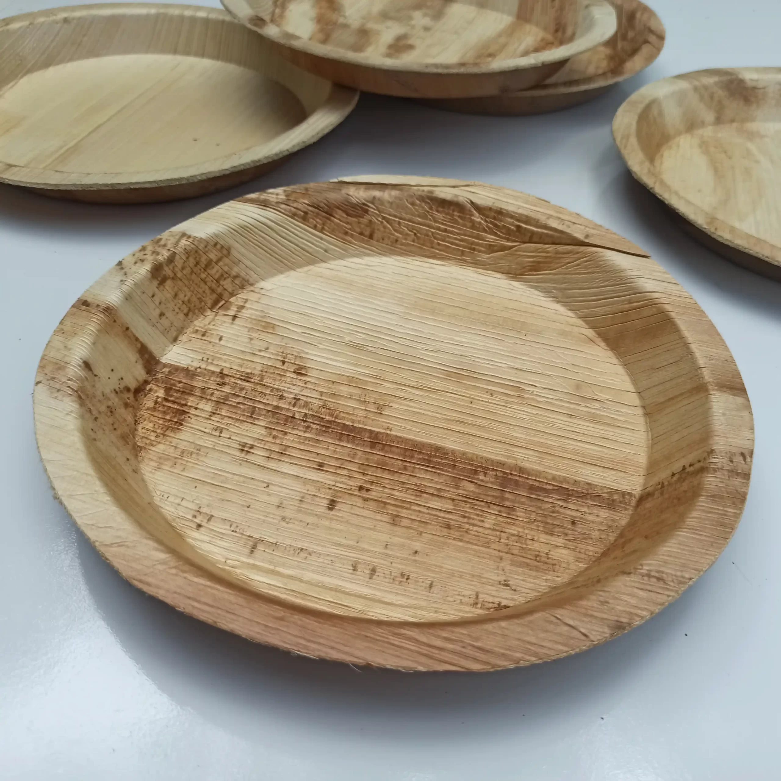 A close-up view of a textured 10 Inch Areca Plate resting on a white table, with several other matching plates in the background.