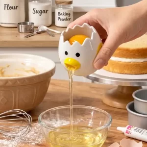 A hand holds a ceramic chicken-shaped egg white separator, which is pouring egg white into a clear glass bowl on a kitchen counter, leaving the yolk inside the separator. Background shows baking ingredients.